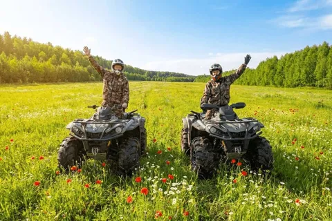 Двое в шлемах и экипировке машут руками, сидя на квадроциклах в поле